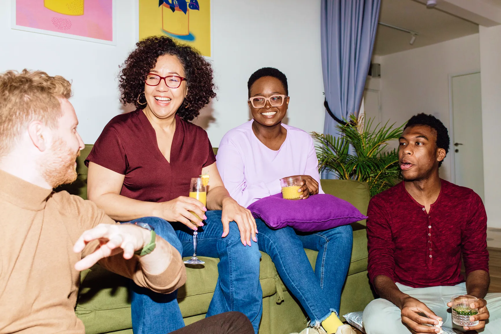 Four diverse friends sitting on a green couch and floor, smiling and holding drinks while chatting indoors.