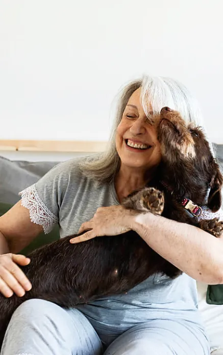 Target customer image: A silver haired woman in 60s sitting on a sofa holding a dog that is about to lick her face